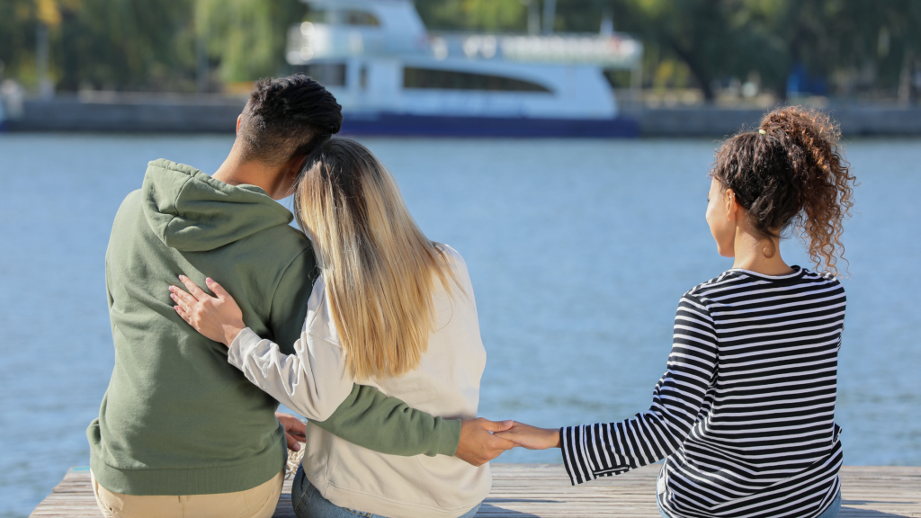 A couple sitting together by the water with one partner looking away, representing discovering that a partner may be involved in a romance scam or online cheating.