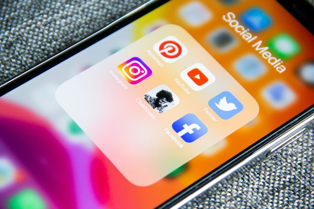 Close-up of an iPhone screen showing a 'Social Media' app folder open, with icons for Instagram, Pinterest, Clubhouse, YouTube, Twitter, and Facebook visible against a blurred orange home screen background