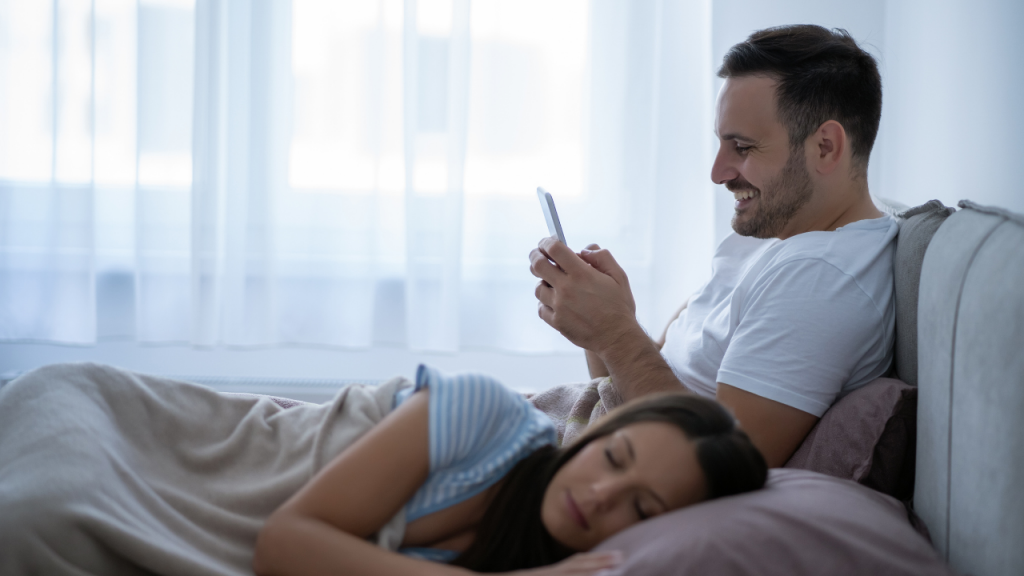 A man texting someone else while his wife is sleeping without her knowing.
