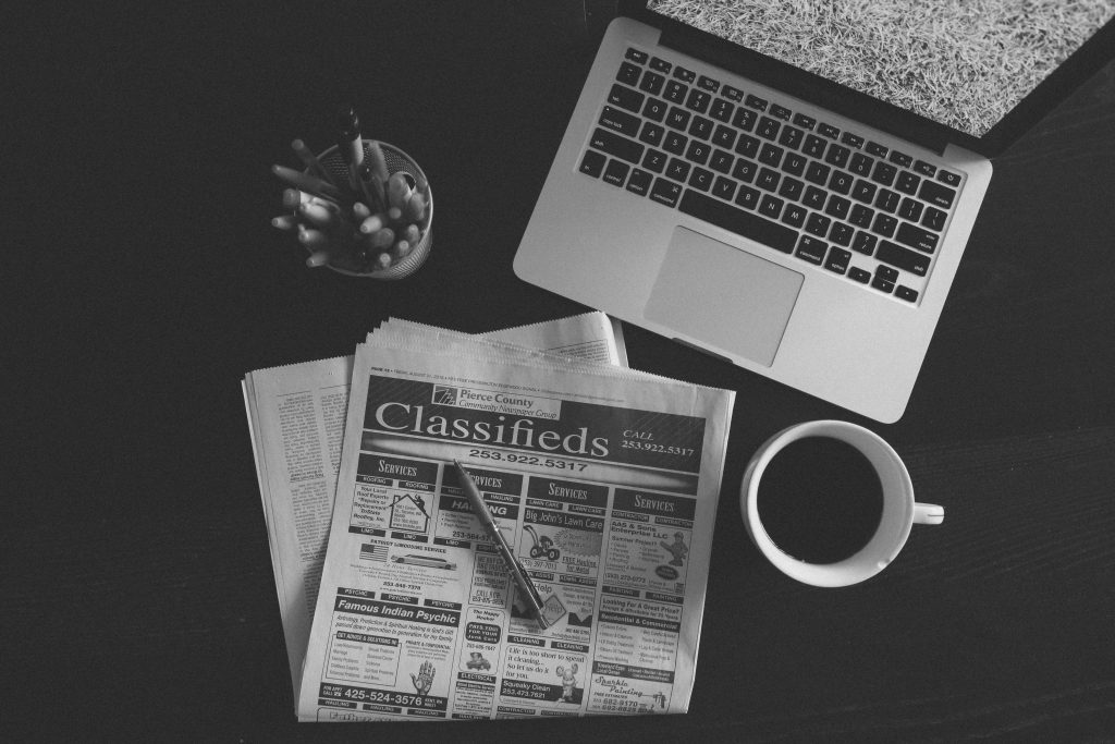 A black and white overhead view of a desk featuring an open laptop, a cup of coffee, a pen holder, and a newspaper open to the local classifieds section.