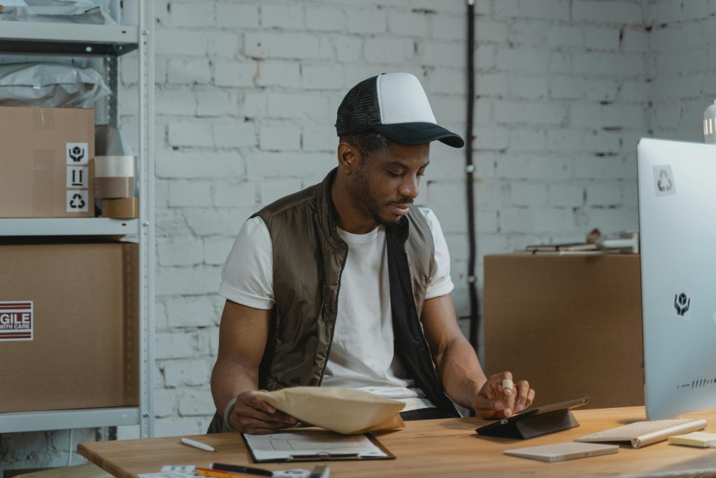 A man wearing a baseball cap and vest sits at a desk in a shipping or warehouse setting, holding a package while looking down at a tablet computer next to shipping boxes.