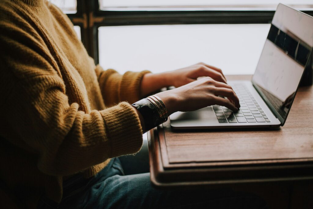 A person in a yellow sweater typing on a laptop by a window, representing searching Tumblr by image to find original photo sources and verify identities online.