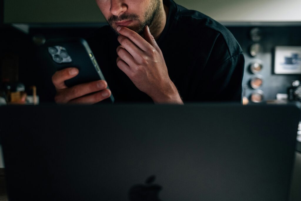 A man looking at his phone with concern in front of a laptop, representing someone discovering identity theft warning signs and checking if someone is using their identity.