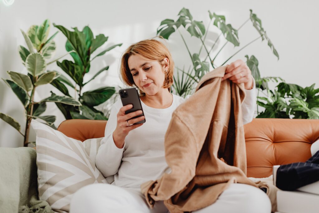 A woman sitting on a couch holding a clothing item while looking at her phone, representing searching Poshmark for secondhand clothing and verifying sellers before making a purchase.
