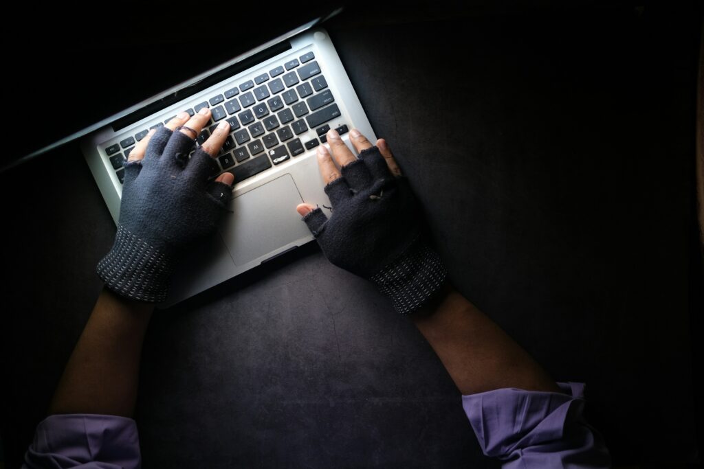 A person wearing fingerless gloves typing on a laptop in the dark, representing identity theft and cybercriminals stealing personal information online.