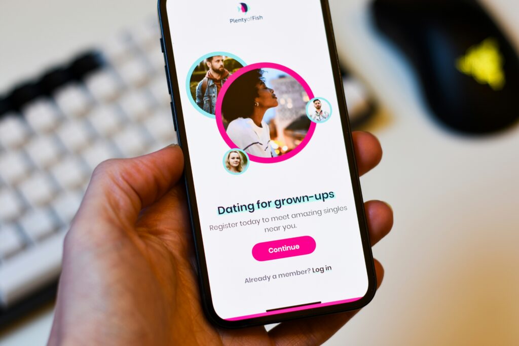 Hand holding a smartphone displaying the Plenty of Fish "Dating for grown-ups" registration screen with member photo circles