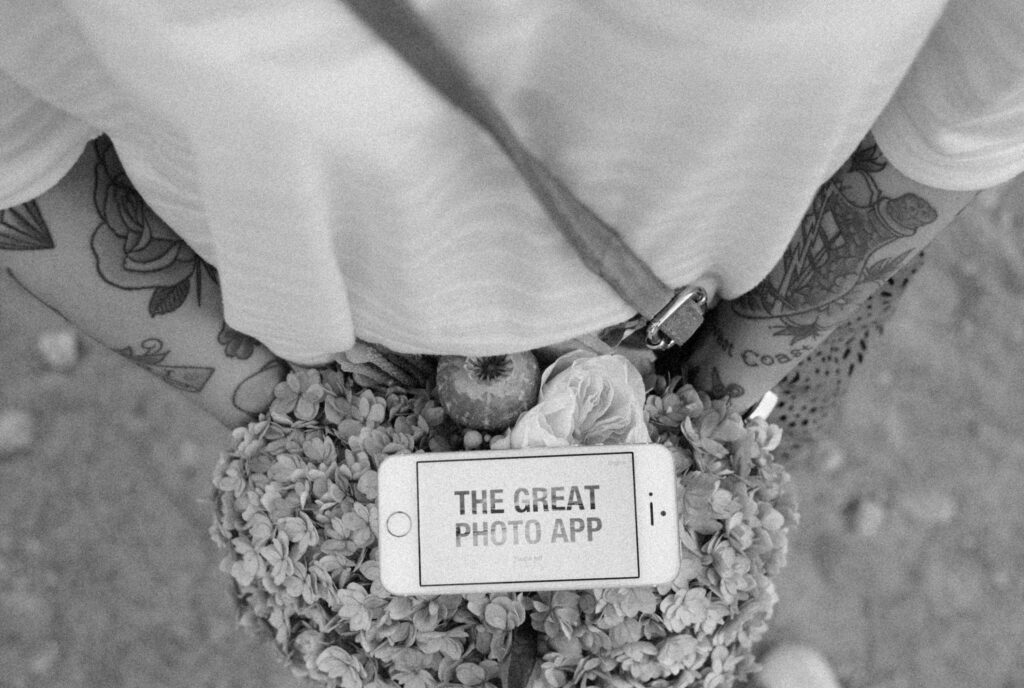 Black-and-white close-up of tattooed legs beside a vintage phone resting on a bed of flowers and a pomegranate, displaying 'The Great Photo App' on its screen.