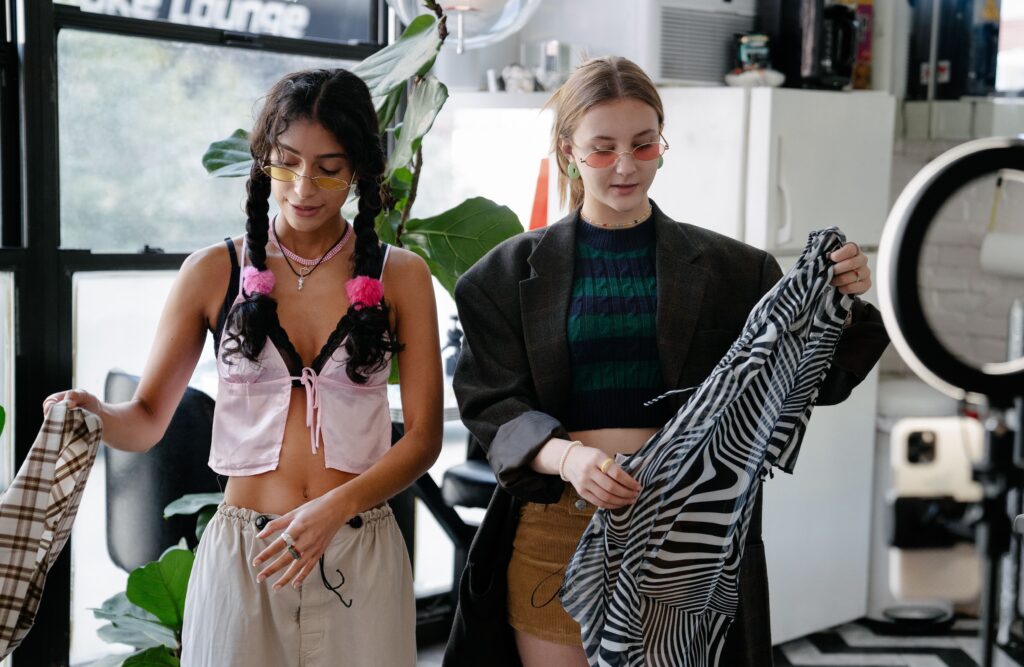 Two young women browsing secondhand clothing items, holding up patterned garments while shopping
