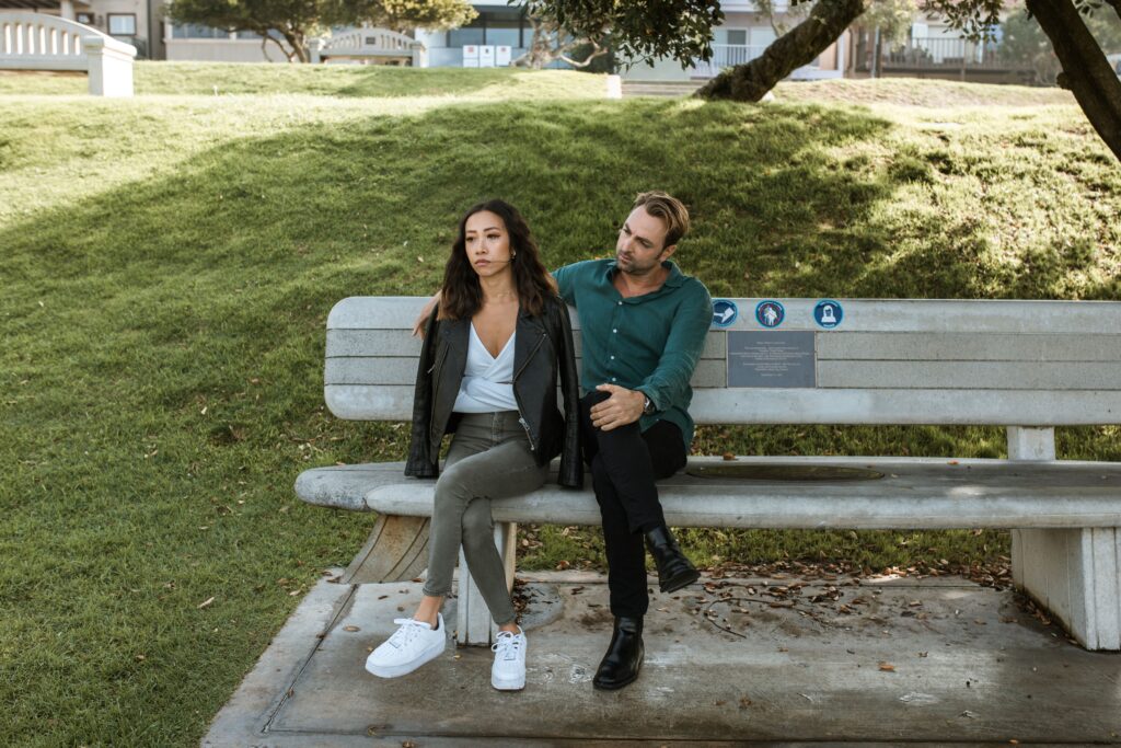 Woman looking away with a distant expression while her partner leans in and talks to her on a park bench.