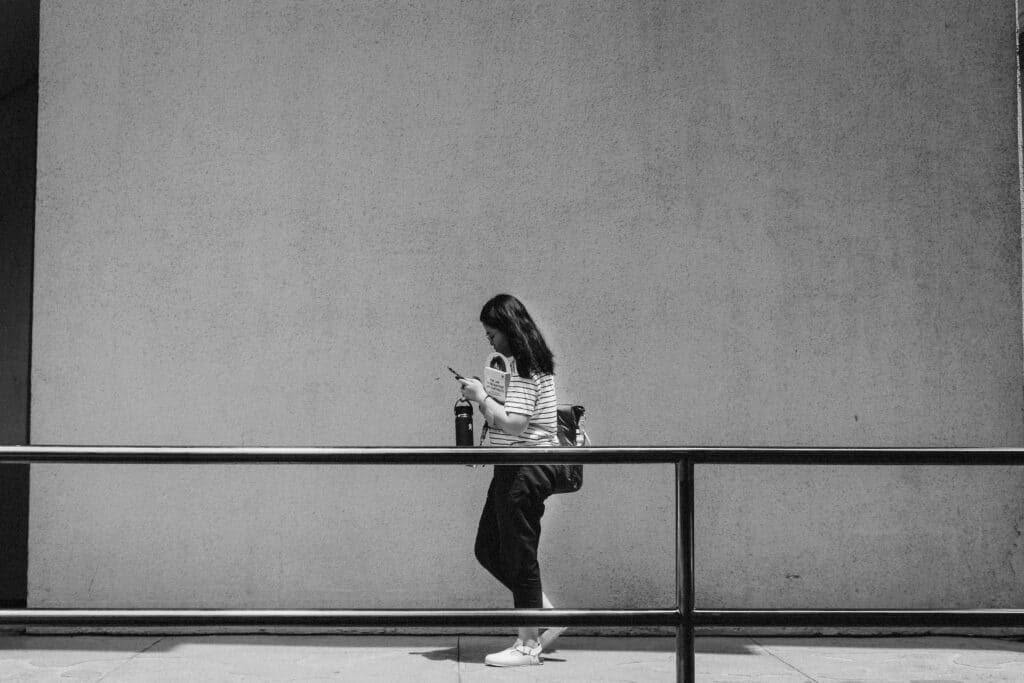 Black and white photo of a young woman leaning against a railing while checking her phone, holding a law textbook