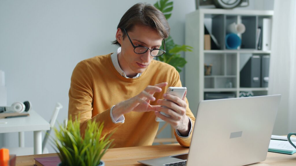 Man in an orange sweater checking his phone next to an open laptop at a home office desk