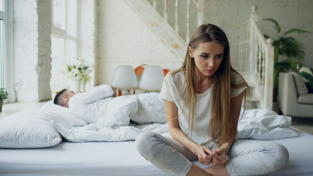 Woman sitting up in bed looking upset and distant while her partner lies turned away from her.