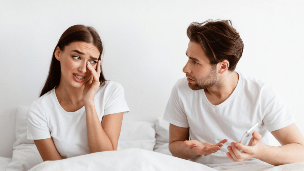 Woman looking upset in bed while a man beside her holds a phone with a questioning expression, illustrating tension over suspected infidelity.