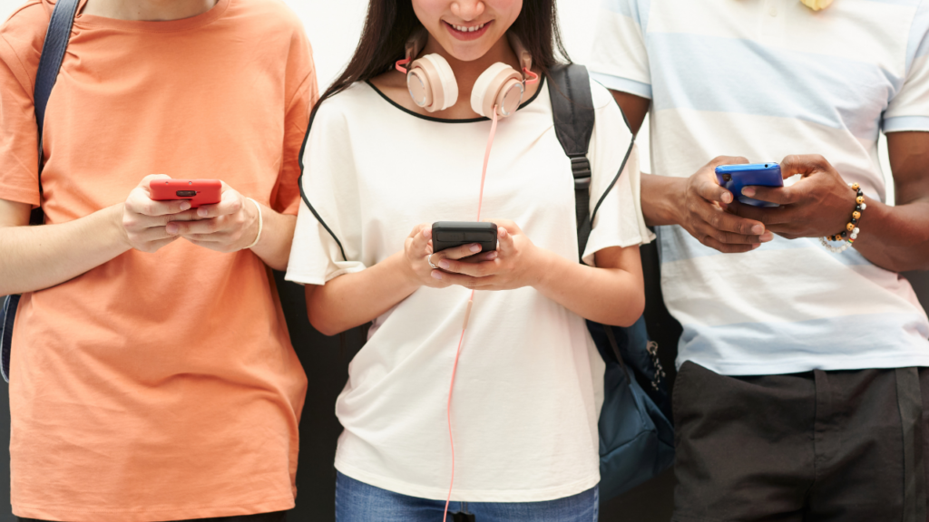 Close-up of three teens standing side by side with backpacks, each looking down at their phone with a smiling girl wearing headphones in the center, reflecting how young users engage with apps like Coverstar.