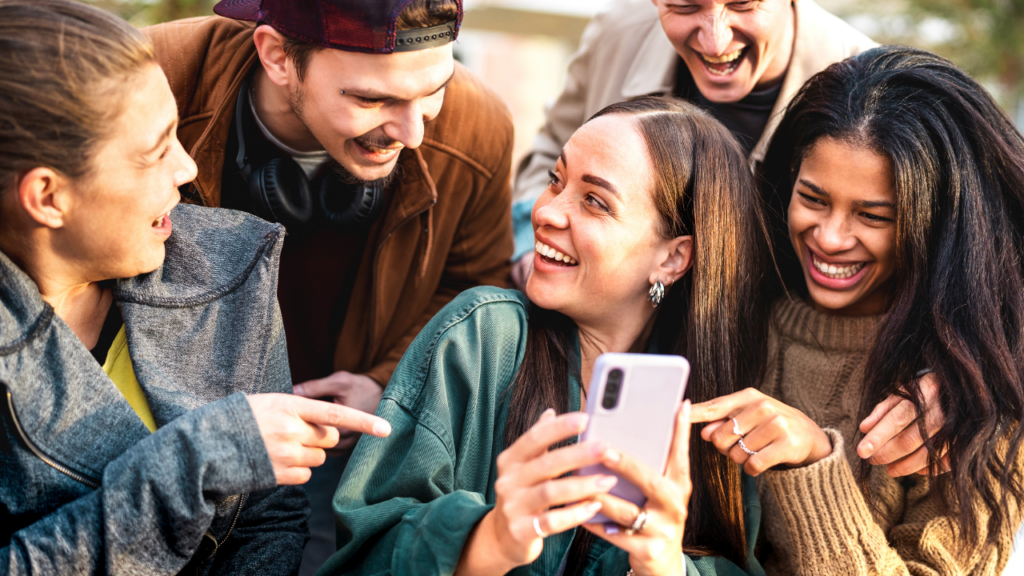 A group of five friends huddled together laughing and pointing at a phone screen, capturing the small-friend-circle photo sharing that apps like Locket are designed around.