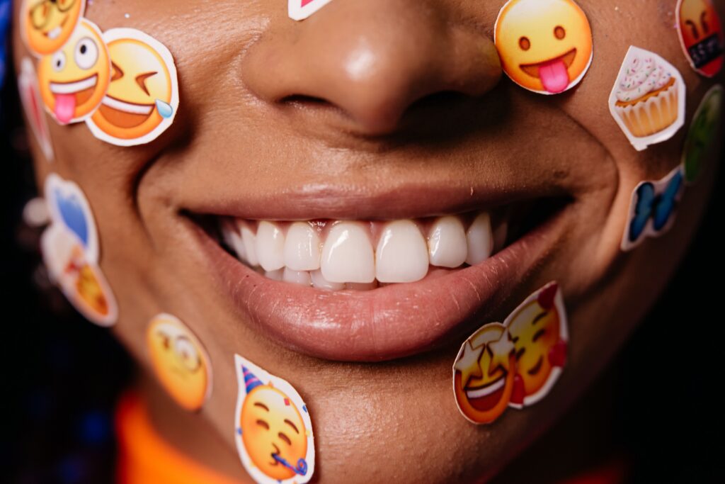 Close-up of a smiling face covered in colorful emoji stickers — including laughing, tongue-out, star-eyes, and party-themed emojis — with a cupcake sticker near the cheek.