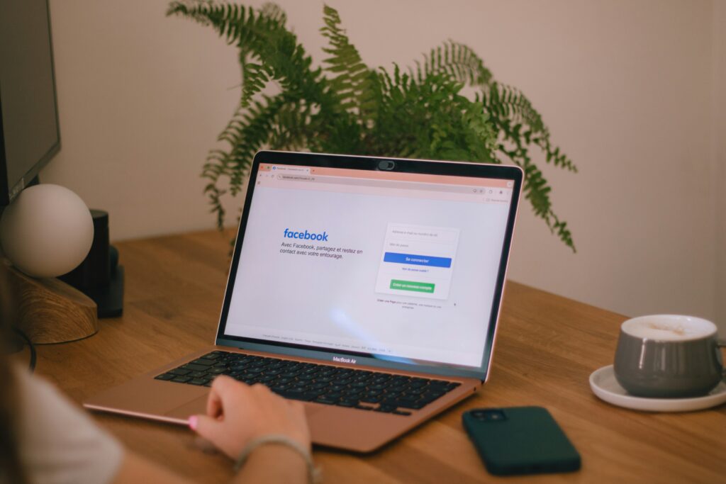 Overhead view of a hand typing on a rose gold MacBook Air displaying the Facebook login page, on a wooden desk with a fern plant, smartphone, and a latte in the background.