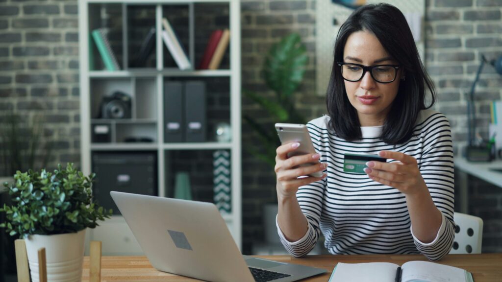 Woman at a desk holding a smartphone and credit card while working on a laptop, illustrating online payment or account access.